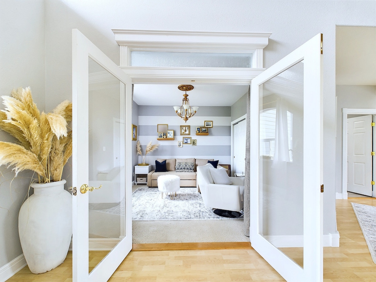 An inviting entryway opens to a bright living area adorned with light-colored walls and decorative wall frames. A plush light-colored rug lies beneath a circular coffee table, and contrasting navy blue pillows are arranged on a soft sofa. A potted plant adds a touch of greenery.