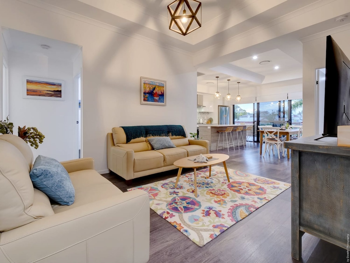 A bright living area features two comfortable sofas and a decorative rug, creating a welcoming space. Natural light flows through large windows, illuminating the open-plan layout that connects to a dining space with a wooden table and modern lighting fixtures.