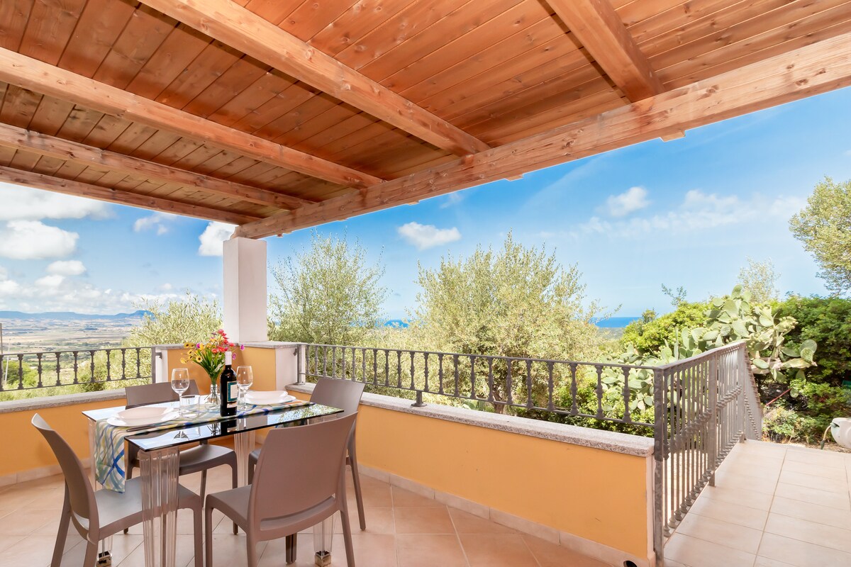 An outdoor terrace is featured with a wooden ceiling and tiled flooring. A dining table is set with glasses and a bottle, surrounded by several grey chairs. Lush greenery frames the partial sea view, which is highlighted against a bright sky.