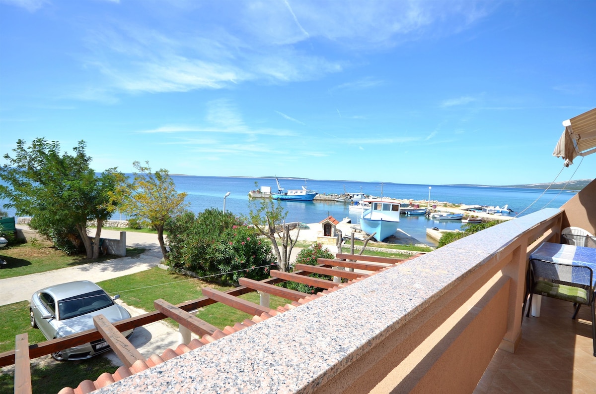 A wide balcony offers a view of the serene sea and nearby marina, featuring boats docked peacefully. Lush greenery and a parking area are visible below, complementing the tranquil atmosphere of the coastal setting.