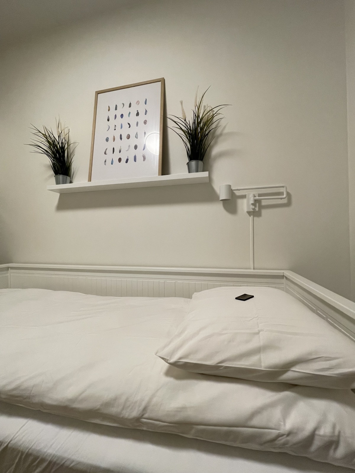 A cozy sleeping area features a spacious bed with white linens and a plump pillow. A shelf above holds two decorative plants and a framed wall art piece. Soft lighting is provided by a wall-mounted lamp, enhancing the calm atmosphere.