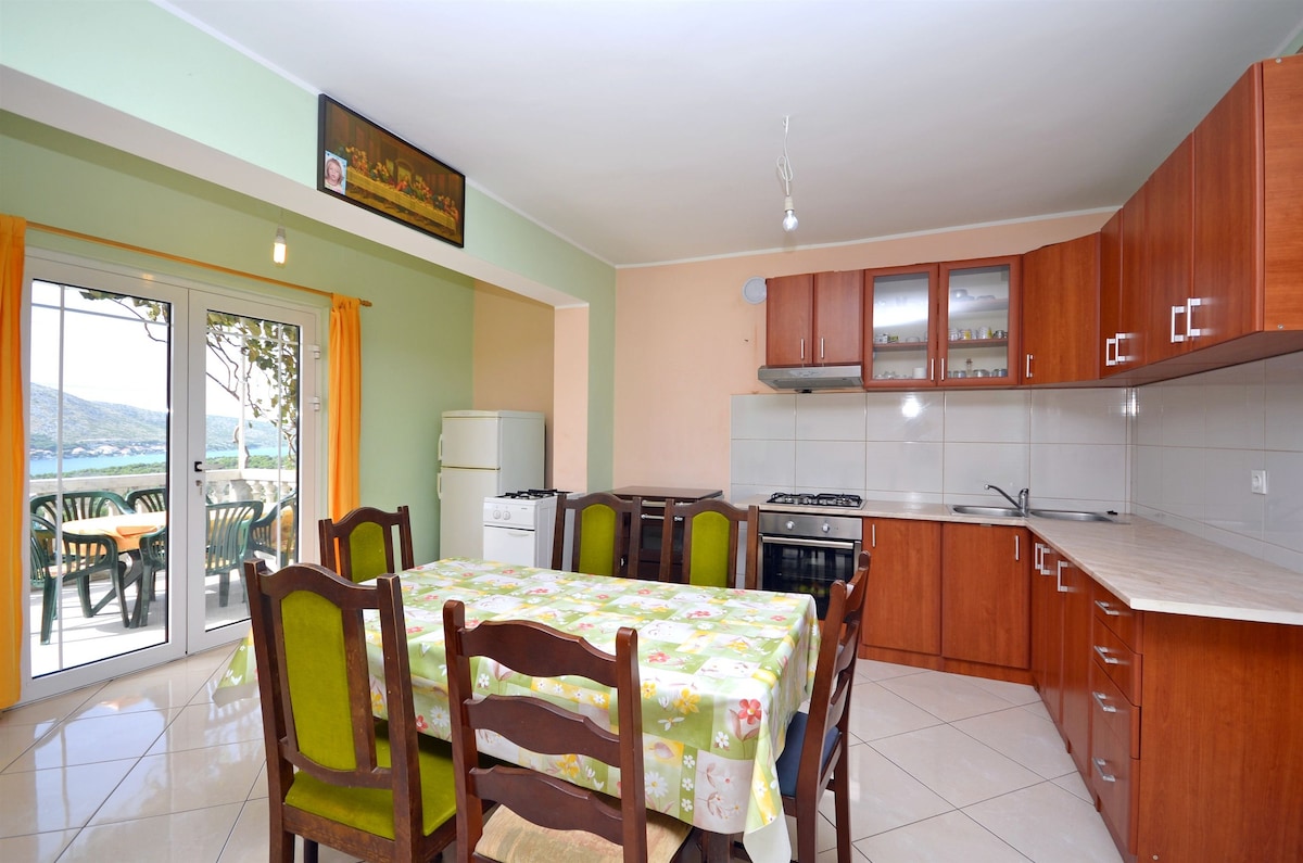 A spacious kitchen area features wooden cabinetry and a large dining table surrounded by chairs. Light-colored tiles cover the floor, and natural light enters through a set of glass doors leading to a terrace. Essential kitchen appliances are visible, ensuring convenience for meal preparation.