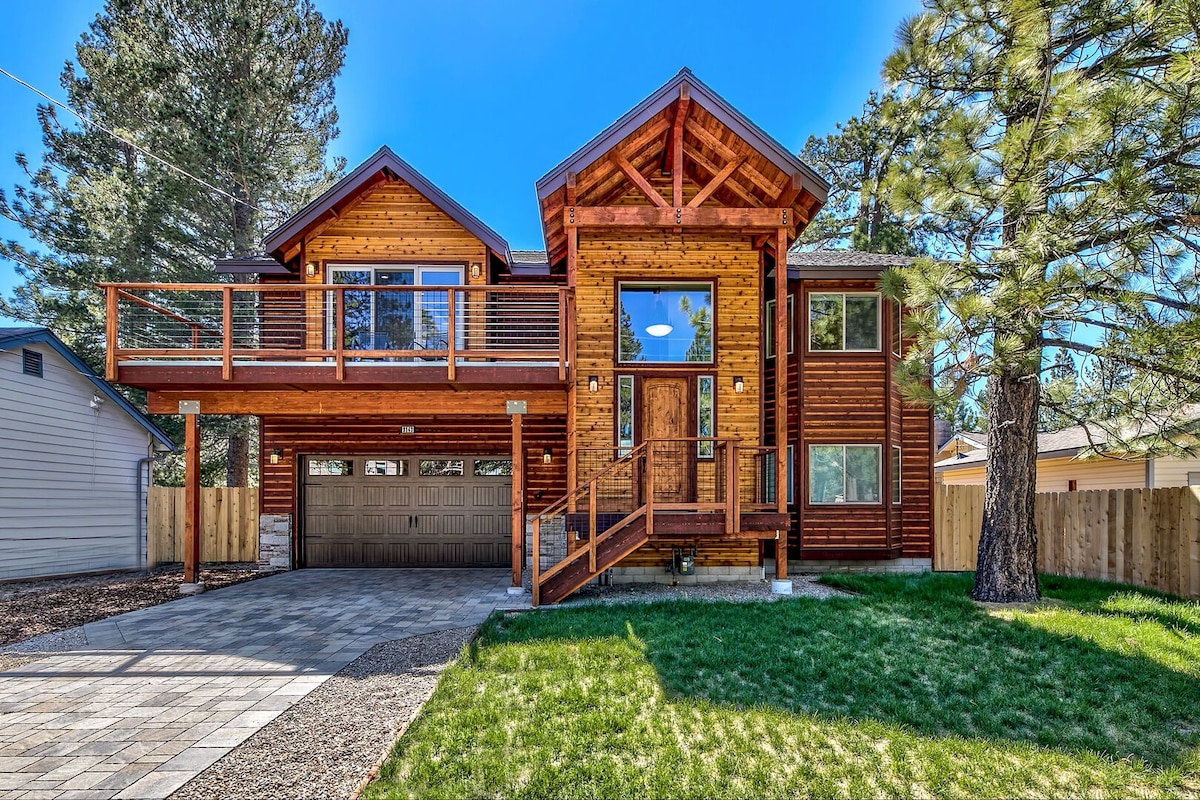 A modern two-story home features a rustic wooden exterior and a spacious front deck. Large windows allow natural light to enter, revealing a welcoming entrance. The well-maintained lawn and surrounding trees enhance the outdoor appeal of the property.