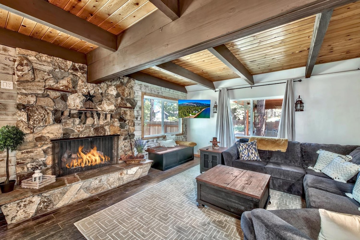 A spacious living area features exposed beam ceilings and a rustic stone fireplace with a warm fire. Comfortable seating is arranged around a wooden coffee table. Large windows provide natural light and views of the outdoor space, which is partially visible through the curtains.