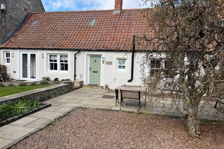 A charming cottage exterior features a red-tiled roof and whitewashed walls. A green door opens into the cozy interior. Stone paving leads to a small table, surrounded by a landscaped garden area with shrubs and a gravel pathway.