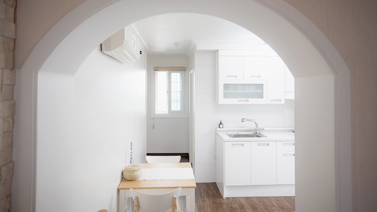 A well-lit kitchen space is visible through an arched entrance. It features modern white cabinets and a sleek sink, complemented by a wooden dining table positioned nearby. Natural light streams in from a window, enhancing the clean and minimalist design.