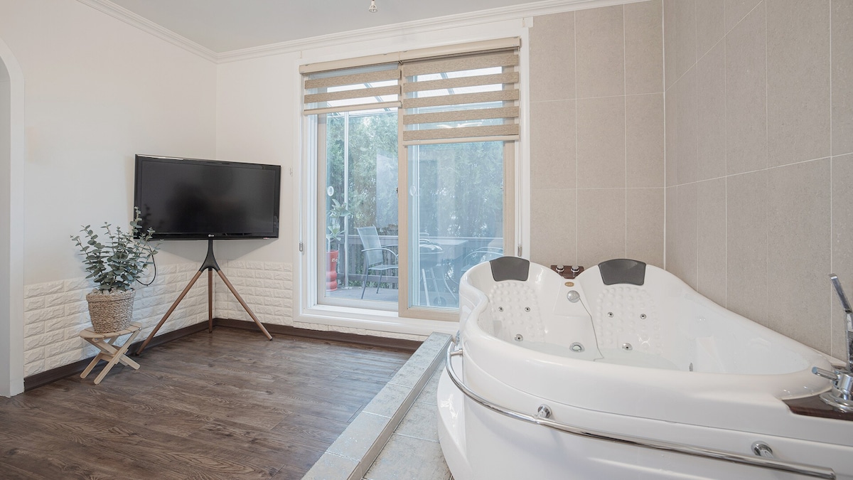 A spacious room features a large spa tub beside a window, offering natural light. A television is mounted on the wall, and wooden flooring is visible throughout the area. Subtle decorations include a small plant on a stand.