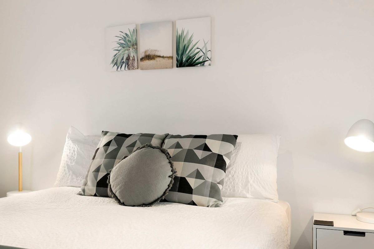 A queen size bed is centered against a white wall, adorned with a geometric-patterned throw pillow. On the wall above, three framed images capture serene nature scenes featuring plants and sandy landscapes. Soft, warm light is emitted from two bedside lamps on either side of the bed.
