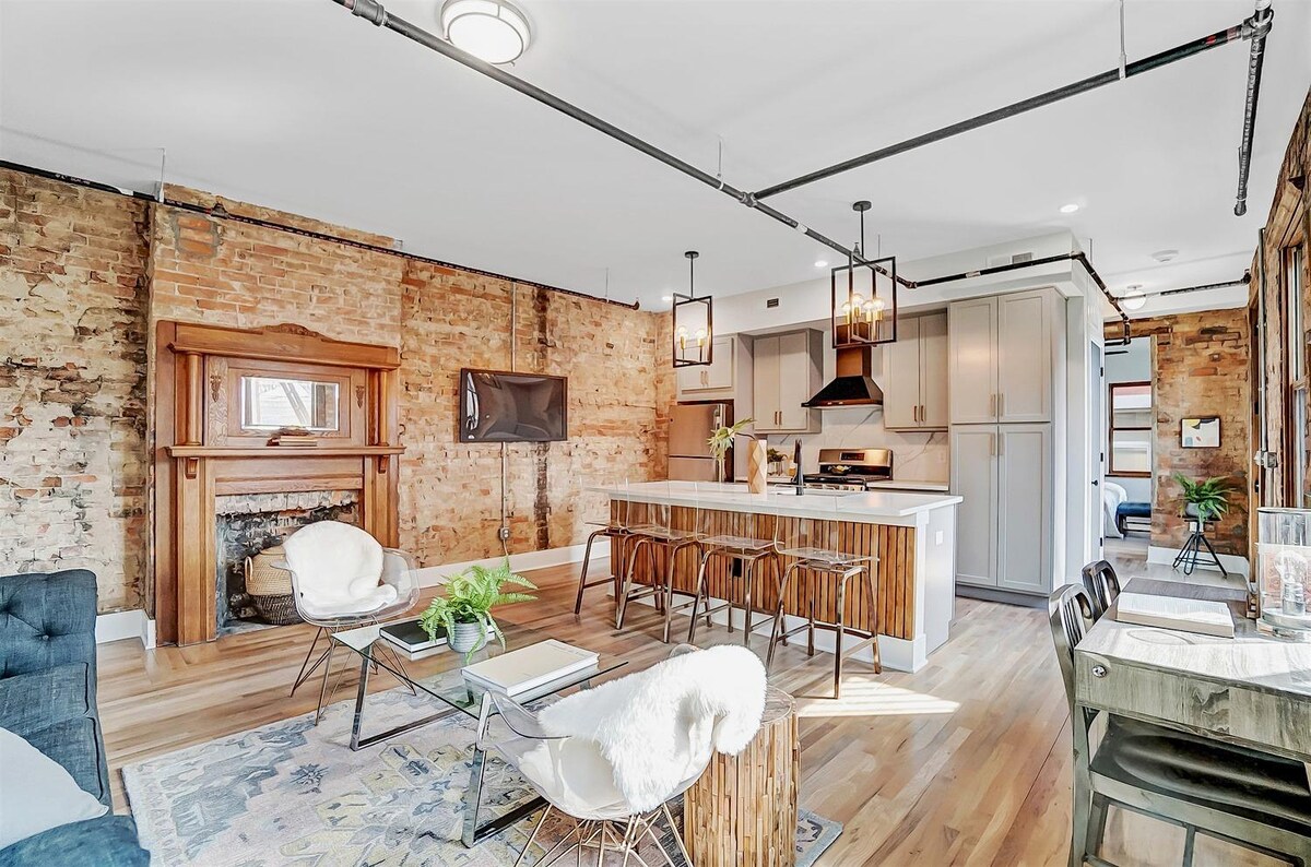 The open-concept living area features exposed brick walls, enhancing the historic charm. A spacious kitchen island with bar seating and stainless steel appliances is visible. Modern furniture and decor create a comfortable space for relaxation and entertainment, while a fireplace mantel adds character.
