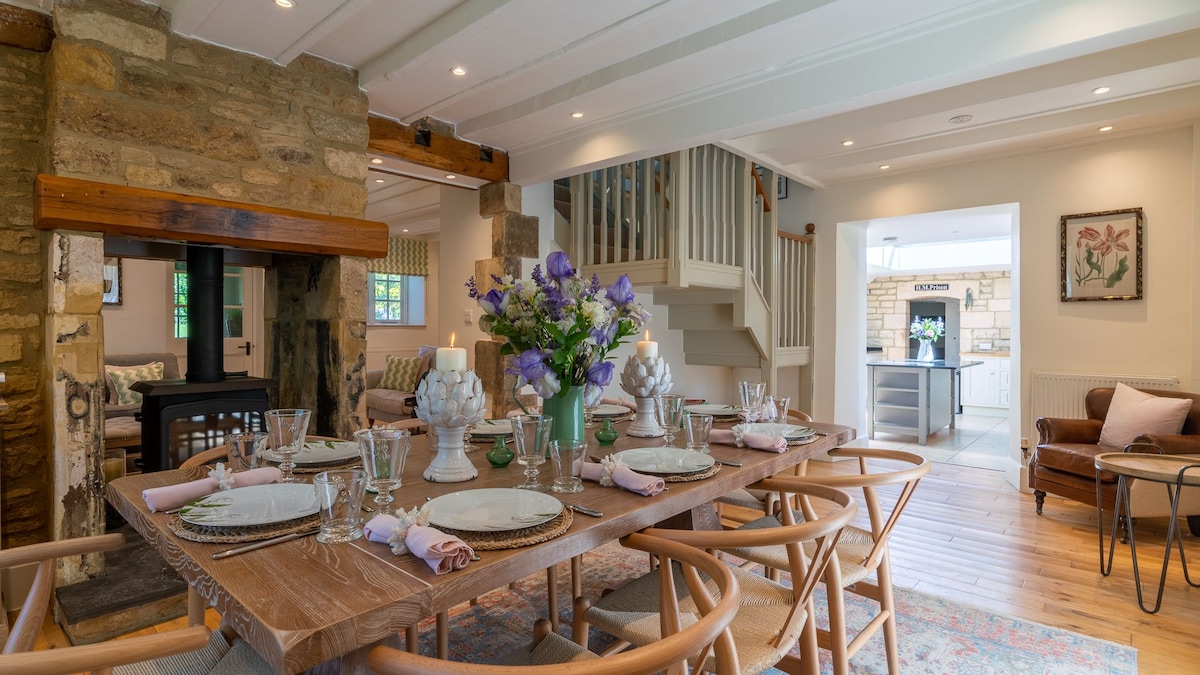 The dining area features a large wooden table set for eight, adorned with elegant tableware and a floral centerpiece. Natural light filters through windows, illuminating the polished wooden floors and stone fireplace. In the background, the open-plan design leads to the kitchen and a charming staircase.