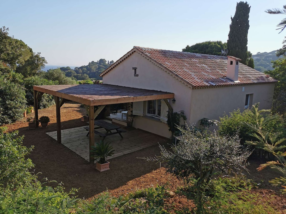 The exterior of a villa is captured, showcasing a covered terrace with wooden beams and a spacious table beneath. The landscape features trees and shrubs, creating a natural setting with a view of the hills in the background. A sense of tranquility is conveyed through the surrounding greenery.