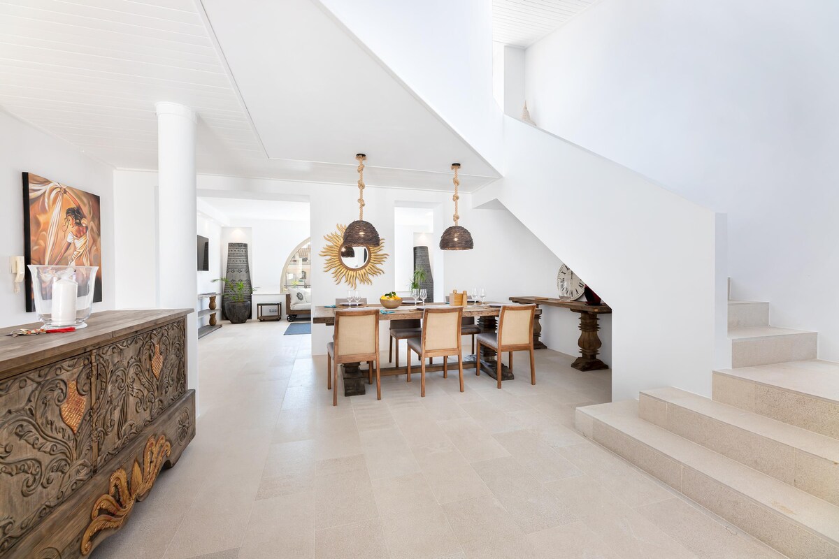 An open and airy living space showcases a dining area with a wooden table and beige chairs underneath pendant lighting. A decorative mirror is displayed on the wall, while stairs lead to the upper level. The floor features light-colored tiles, enhancing the bright atmosphere.