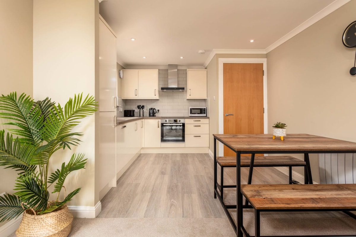 A modern kitchen space is complemented by light-colored cabinets and stainless steel appliances. A wooden dining table with black metal legs accommodates seating for four. The floor is finished with a light wood tone, enhancing the open and airy feel of the area.