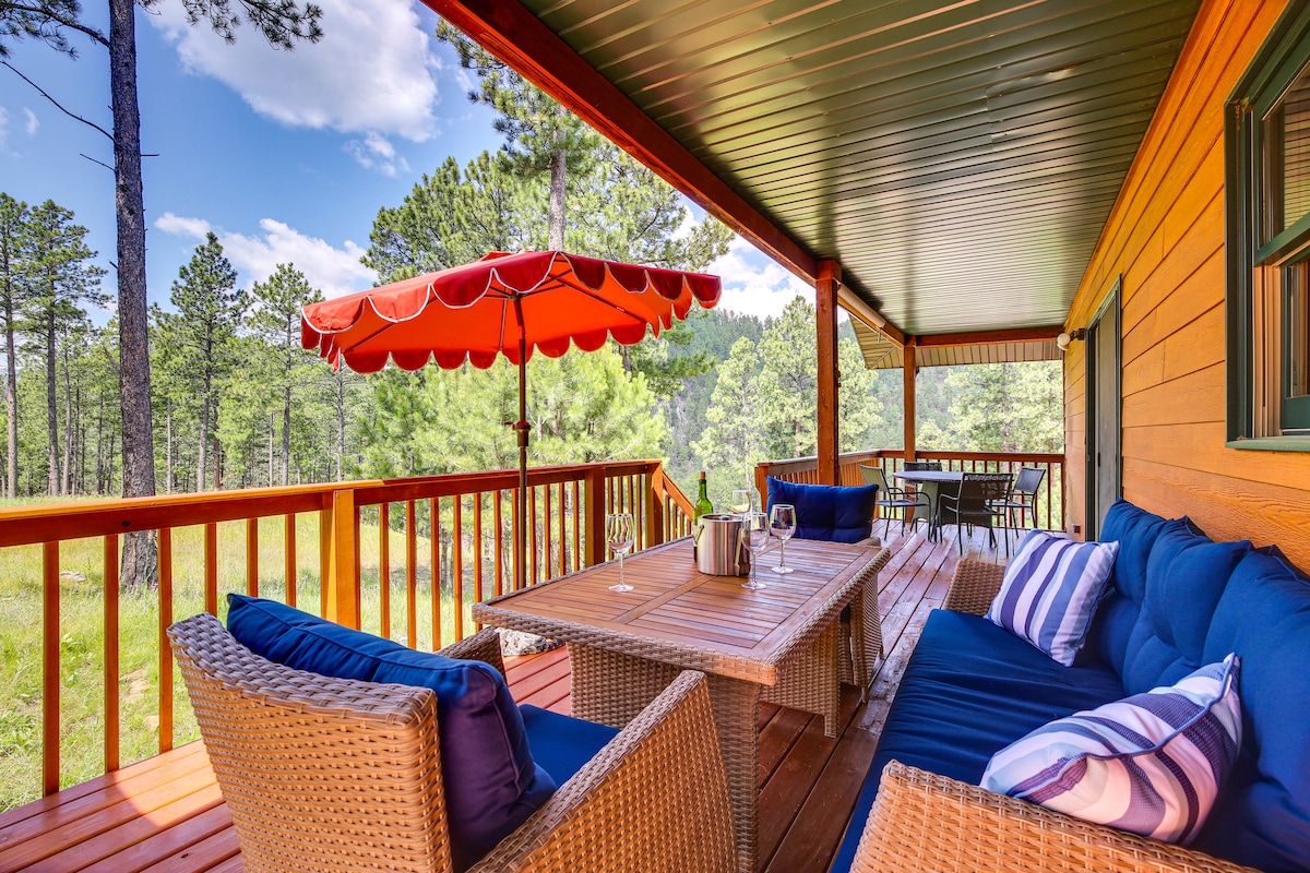 A spacious outdoor deck is furnished with comfortable seating and a dining table, surrounded by natural greenery. An umbrella provides shade, while views of the surrounding trees can be seen in the background under a partly cloudy sky.
