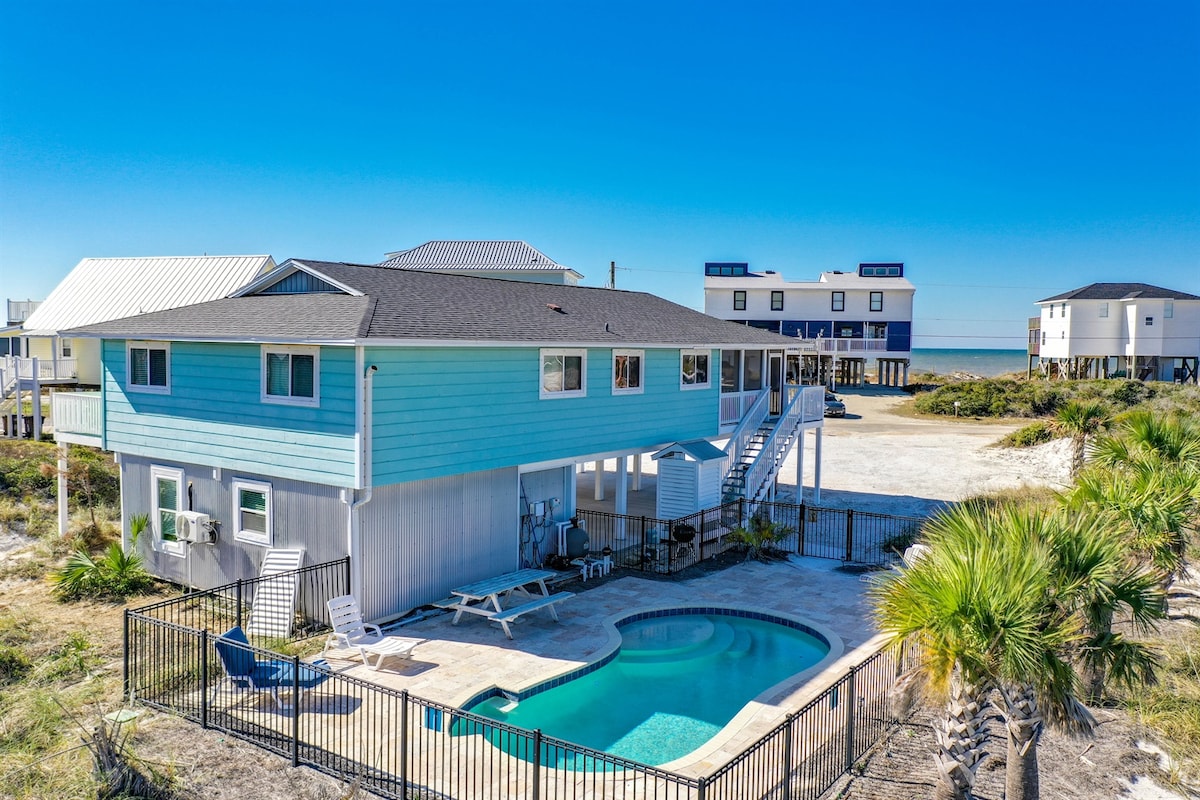 A light blue house is featured, set against a clear blue sky. A private pool is surrounded by a fenced patio area, with lounge chairs positioned nearby. The sandy area extends towards several wooden structures in the background, indicating a coastal location.