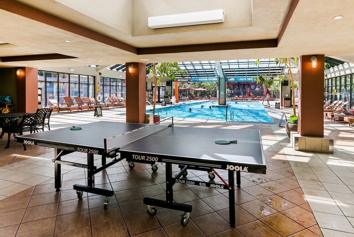 Two ping pong tables are positioned in a spacious indoor area overlooking a large swimming pool. Natural light floods the space through a glass ceiling, illuminating the tiled floor and the chairs arranged around the pool. Several guests are seen enjoying the pool in the background.