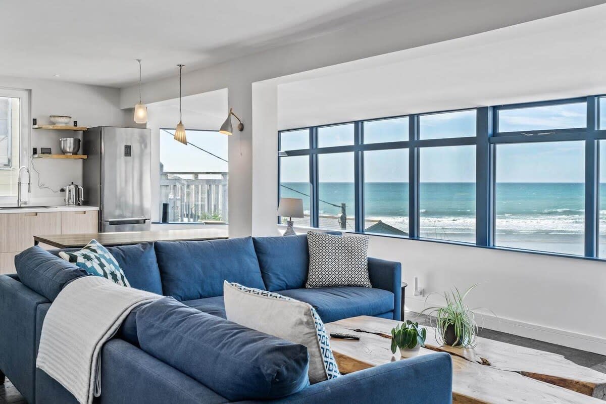 A spacious living area is designed with a blue corner sofa, facing large windows that offer panoramic sea views. A wooden coffee table sits at the center, complemented by decorative plants. The light-filled space features modern kitchen appliances visible in the background.
