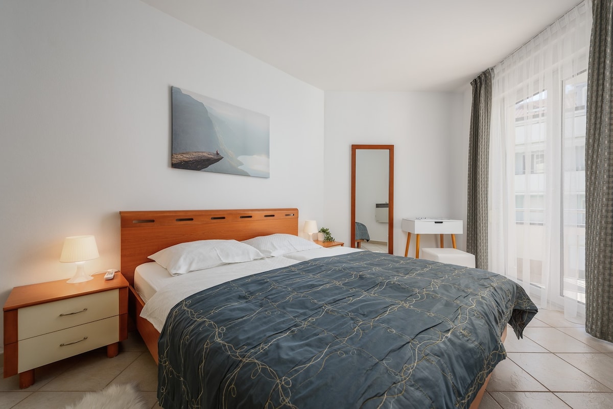 A bright bedroom features a spacious bed with a dark blue coverlet. A large window with sheer curtains allows natural light to fill the room. A mirror and a bedside table are positioned beside the bed, enhancing the room's functionality.