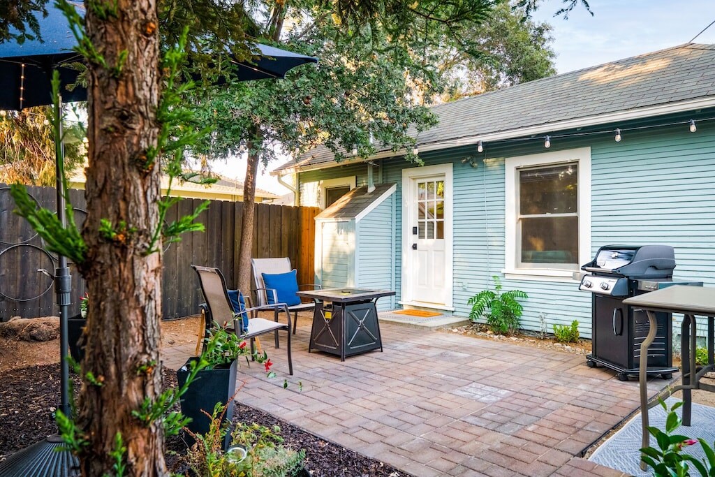 A private yard features a smooth brick patio surrounded by greenery. A grill is positioned next to a dining table and chairs. Soft bistro lighting is strung overhead, enhancing the comfortable seating area with a coffee table and two lounge chairs.