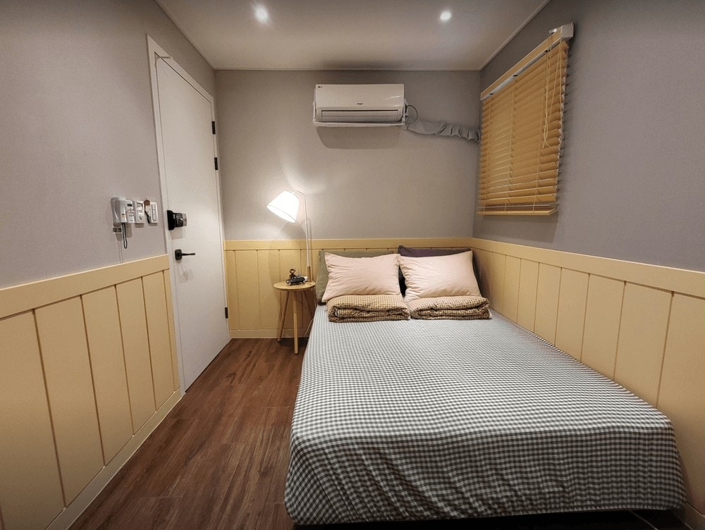The room features a compact sleeping area with a double bed dressed in light bedding. A bedside table holds a lamp, and natural light enters through a window covered by wooden blinds. Soft wall colors and wooden accents provide a calming effect.