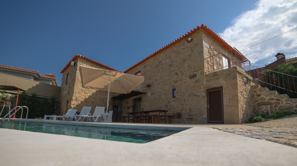 A rustic vacation house is presented with stone exterior and terracotta roof under a clear blue sky. The private pool is surrounded by sun loungers, while a shaded outdoor dining area is visible. The overall setting offers an inviting space for relaxation and enjoyment.