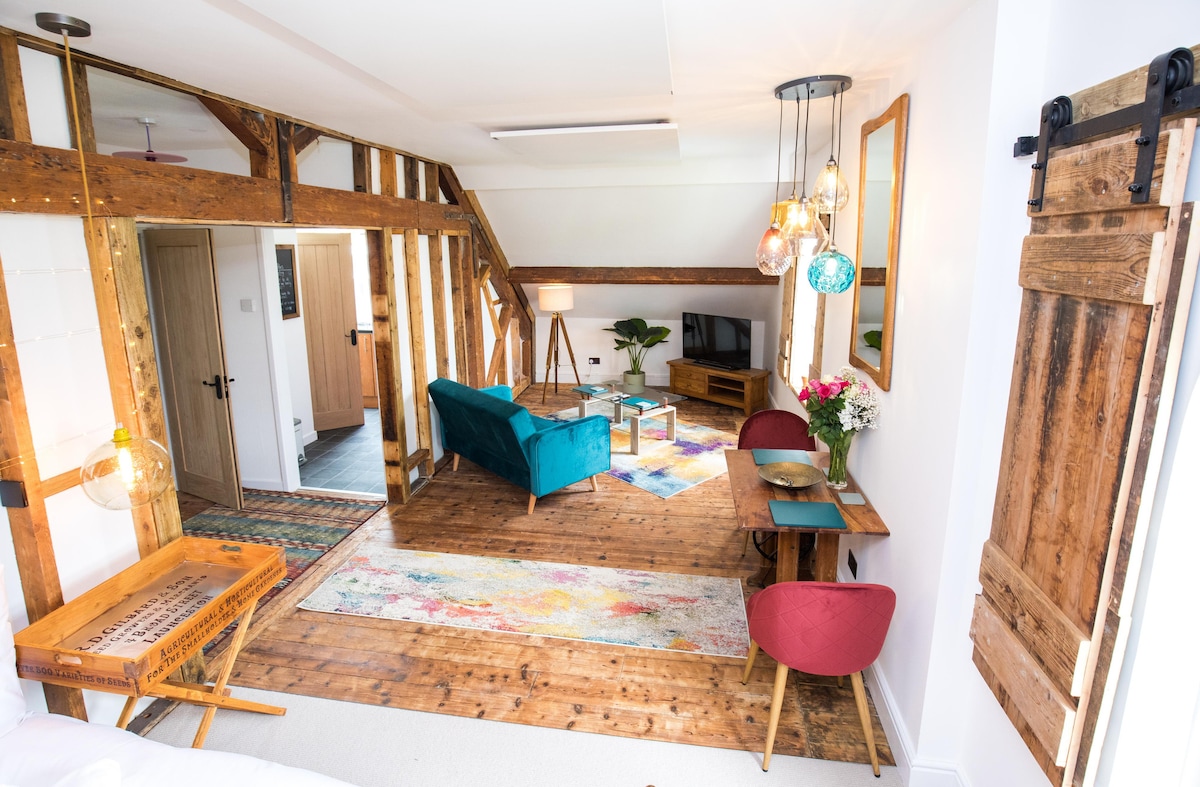 An open-plan studio is showcased, featuring exposed timber beams and vintage floorboards. A teal velvet sofa and red velvet chairs provide seating, while a small bistro table and Smart TV are present. Decorative pendant lights hang from the ceiling, enhancing the space.