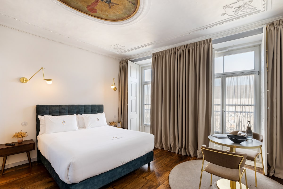 A spacious bedroom features a large bed with a dark green velvet headboard. Natural light enters through two windows covered by flowing curtains. A small round table with chairs and a decorative rug adds an inviting touch, while decorative ceiling details enhance the historic ambiance.