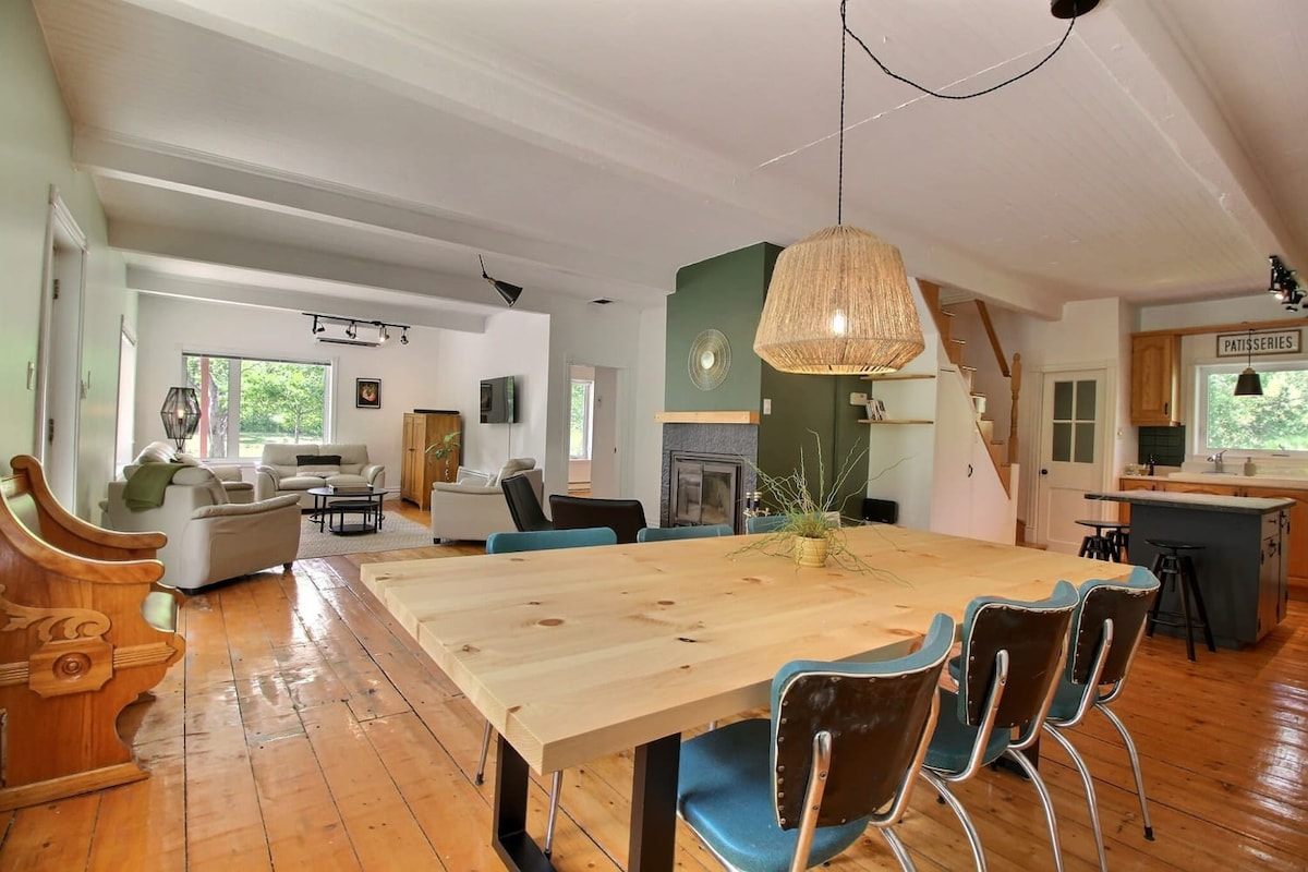 An open-concept living and dining area showcases a large wooden table surrounded by seating. Soft seating arrangements are visible in the background, along with a fireplace. Natural light floods the space through the windows, providing a welcoming ambiance.