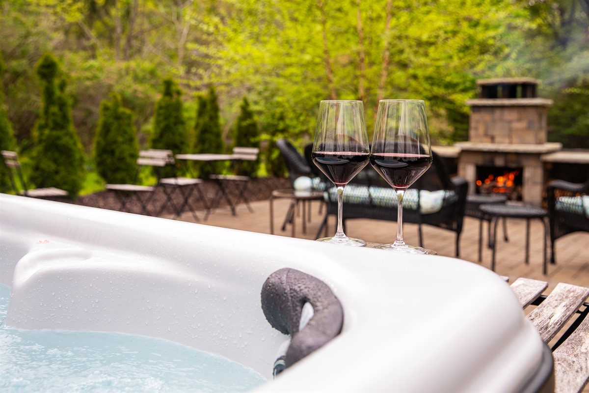 A hot tub is shown with bubbling water, accompanied by two glasses of red wine placed on the edge. Surrounding the hot tub, a cozy outdoor space features a stone fireplace and seating arrangements including small tables and chairs nestled among greenery.