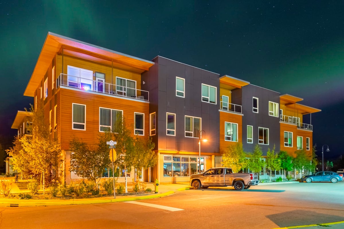 A modern three-story building is illuminated at night, featuring a mix of wooden and dark exterior finishes. Balconies with glass railings are visible on the upper levels, and landscaped greenery frames the entrance. The parking area includes a pickup truck parked nearby.
