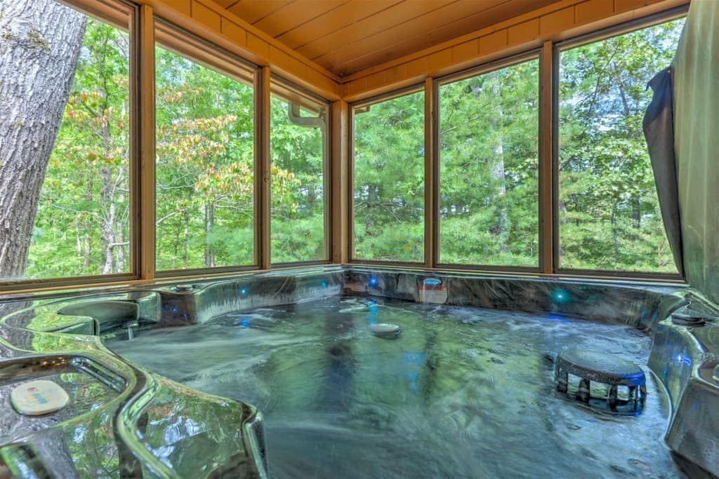 A spacious hot tub is situated within a screened area, surrounded by large windows that provide views of the lush greenery outside. Soft lighting enhances the relaxing atmosphere, creating a tranquil spot for unwinding in nature.