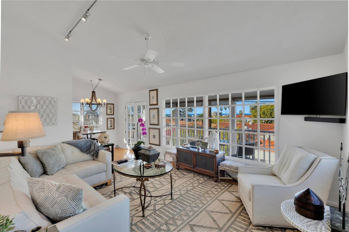 A light-filled living room with large windows offers views of the surrounding area. Comfortable seating arrangements include a spacious sofa and lounge chair. A round glass table is centrally located, accented with decorative pieces. Modern decor enhances the open and airy ambiance.