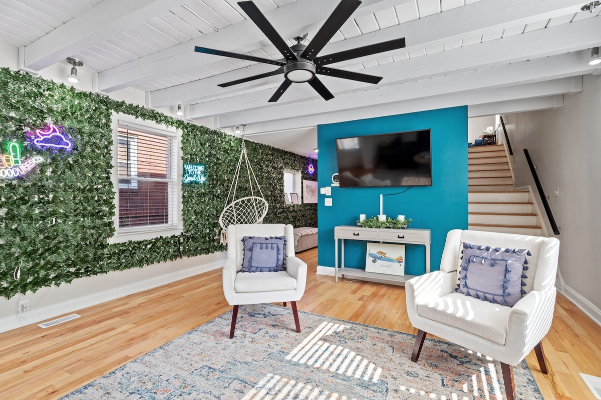A bright living area features two cozy chairs with decorative pillows. A large ceiling fan enhances airflow, while vibrant green wall accents and a bold blue wall add visual interest. A television is mounted on one wall, and stairs lead to the upper level.