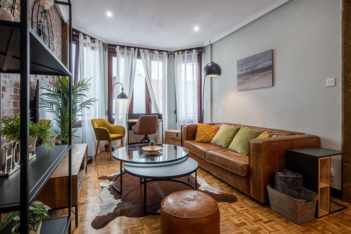 A modern living area features a leather sofa paired with colorful accent chairs. A circular coffee table sits on a patterned rug, while large windows dressed with sheer curtains allow natural light to brighten the space. Decorative shelving units add character without cluttering the room.