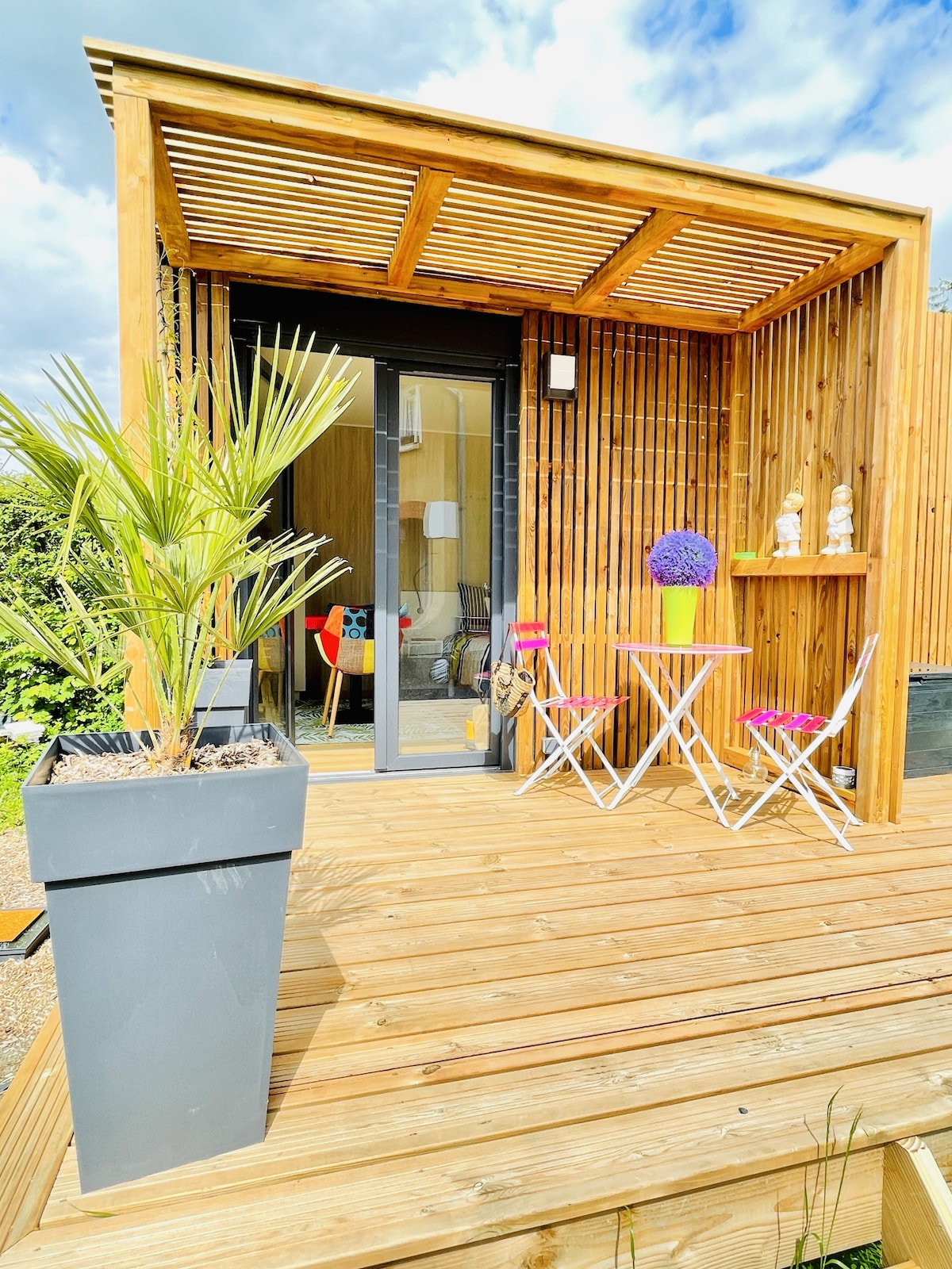 An independent terrace is visible, featuring a wooden deck and a decorative planter. A small table and two chairs are arranged, accompanied by vibrant flower arrangements. Sliding glass doors lead into the interior space, illuminated by natural light, creating a serene outdoor setting.