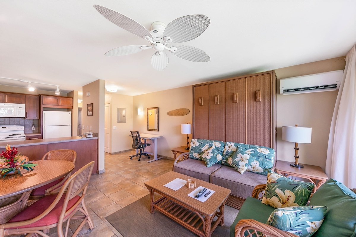 A light-filled living area features a spacious sofa adorned with tropical-patterned cushions. A round dining table accommodates six, and a desk with USB charger ports is visible in the background. Air conditioning is provided, and a ceiling fan enhances air circulation.