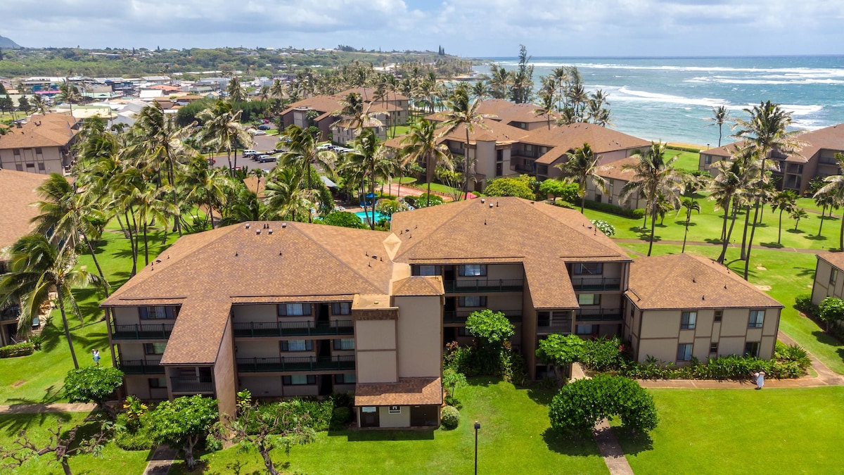 An aerial view of the Pono Kai Resort showcases its well-maintained buildings surrounded by lush greenery and palm trees. The ocean glimmers in the background, and the nearby town can be seen in the distance, creating a picturesque setting.