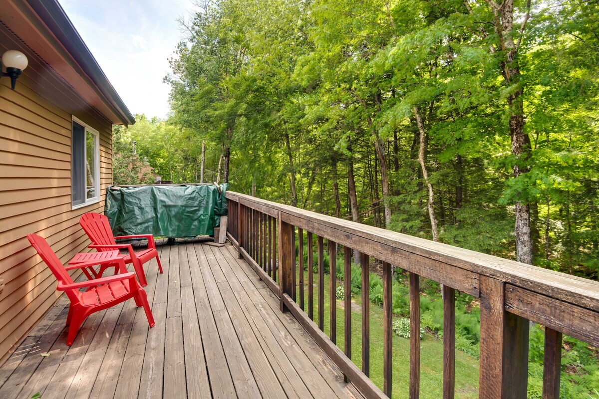 A private balcony features wooden flooring and overlooks a lush, green forest. Two red chairs are positioned beside a covered grill, inviting relaxation and outdoor dining. The surrounding trees create a serene atmosphere, providing a peaceful retreat.