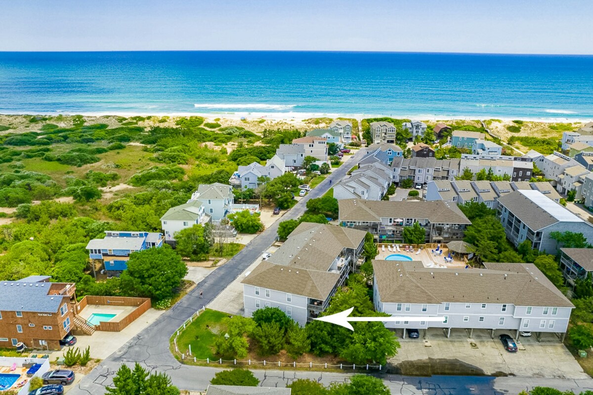An aerial view captures a coastal community, with the beach visible in the background. Multiple buildings are shown, surrounded by greenery, while a shared pool area is highlighted nearby. The ocean's blue waves contrast with the land, emphasizing the proximity to the shoreline.