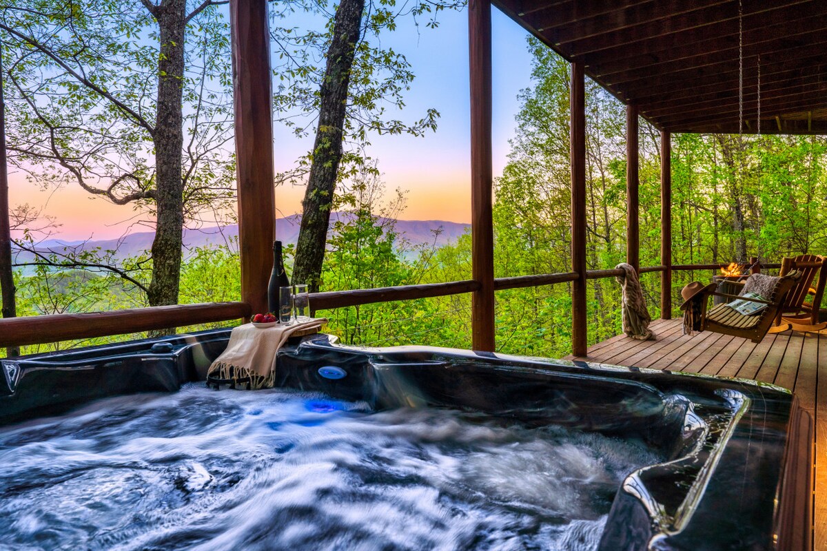 A relaxing hot tub is situated on a wooden deck, surrounded by lush greenery. The view of the mountains is visible in the background, complemented by a soft sunset glow. Two rustic chairs are positioned nearby, inviting quiet moments of enjoyment.