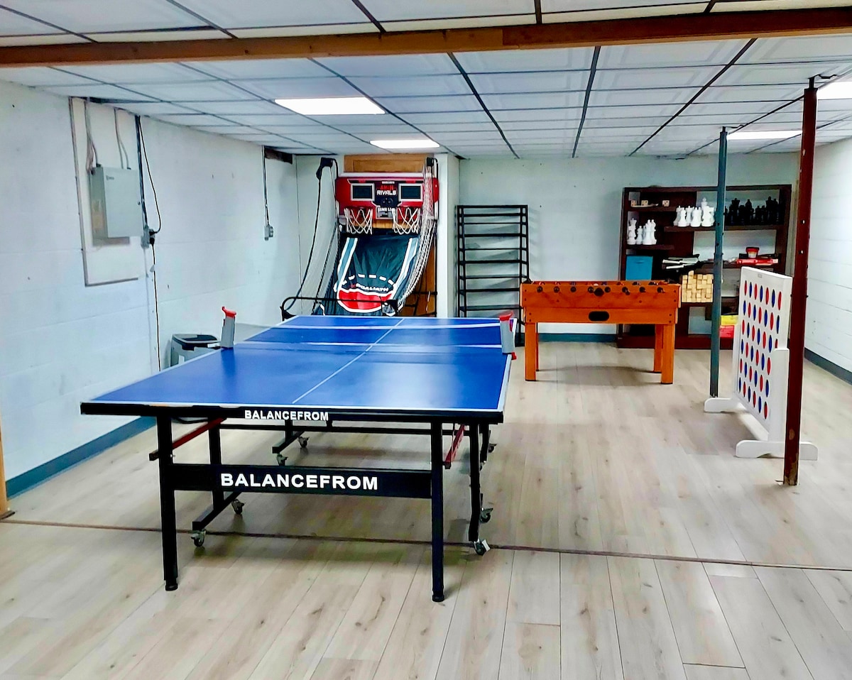 The recreation room features a table tennis setup and a Foosball table, with colorful walls and wood flooring. A basketball shooting game and oversized chess set are present in the background, providing various entertainment options for guests.