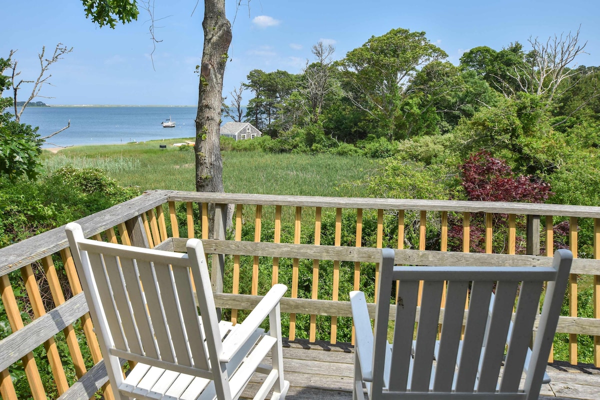 A wooden deck with two white rocking chairs overlooks a serene bay view. Lush greenery and tall grasses frame the scene, while a calm water expanse can be seen in the distance, with a small boat anchored nearby.