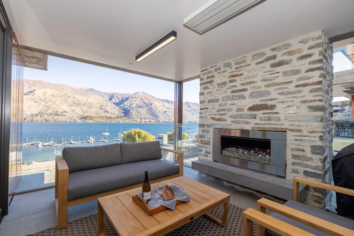 A modern outdoor lounge area features a sleek grey sofa and wooden chairs, positioned around a natural wood table. A stone fireplace adds warmth, with large windows providing panoramic views of the lake and mountains in the background.