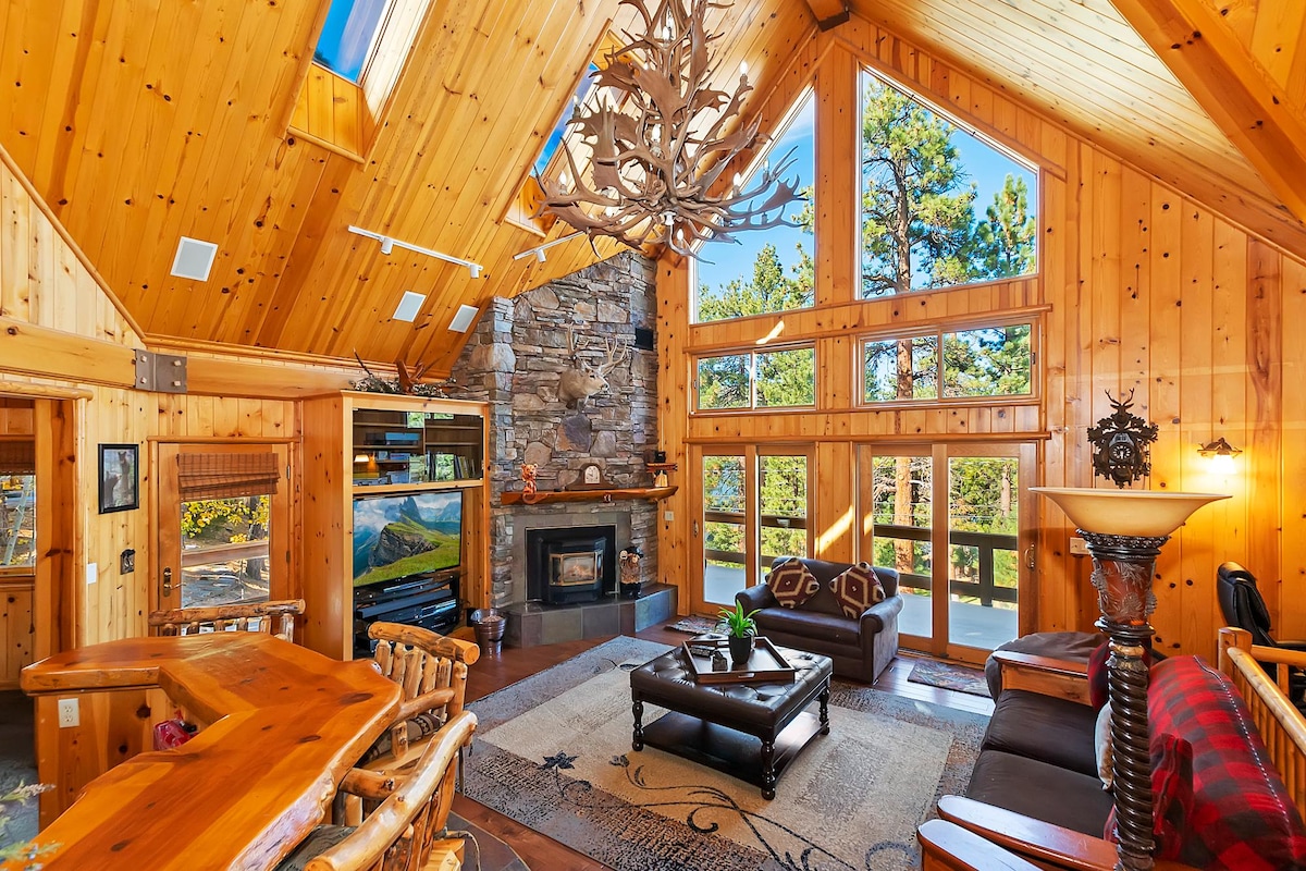 The spacious living area features high vaulted ceilings and floor-to-ceiling windows, allowing natural light to fill the space. A rustic stone fireplace is centered with an antler chandelier above. Cozy seating is arranged around a central coffee table, with wood accents enhancing the warm environment.