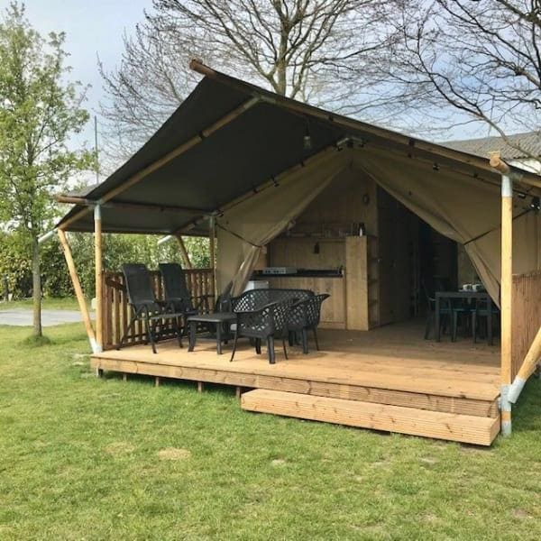 A safari tent features a spacious deck with a wooden floor. The covered area includes several chairs and a table, offering a comfortable space for outdoor dining. Lush greenery surrounds the tent, creating a serene outdoor setting.