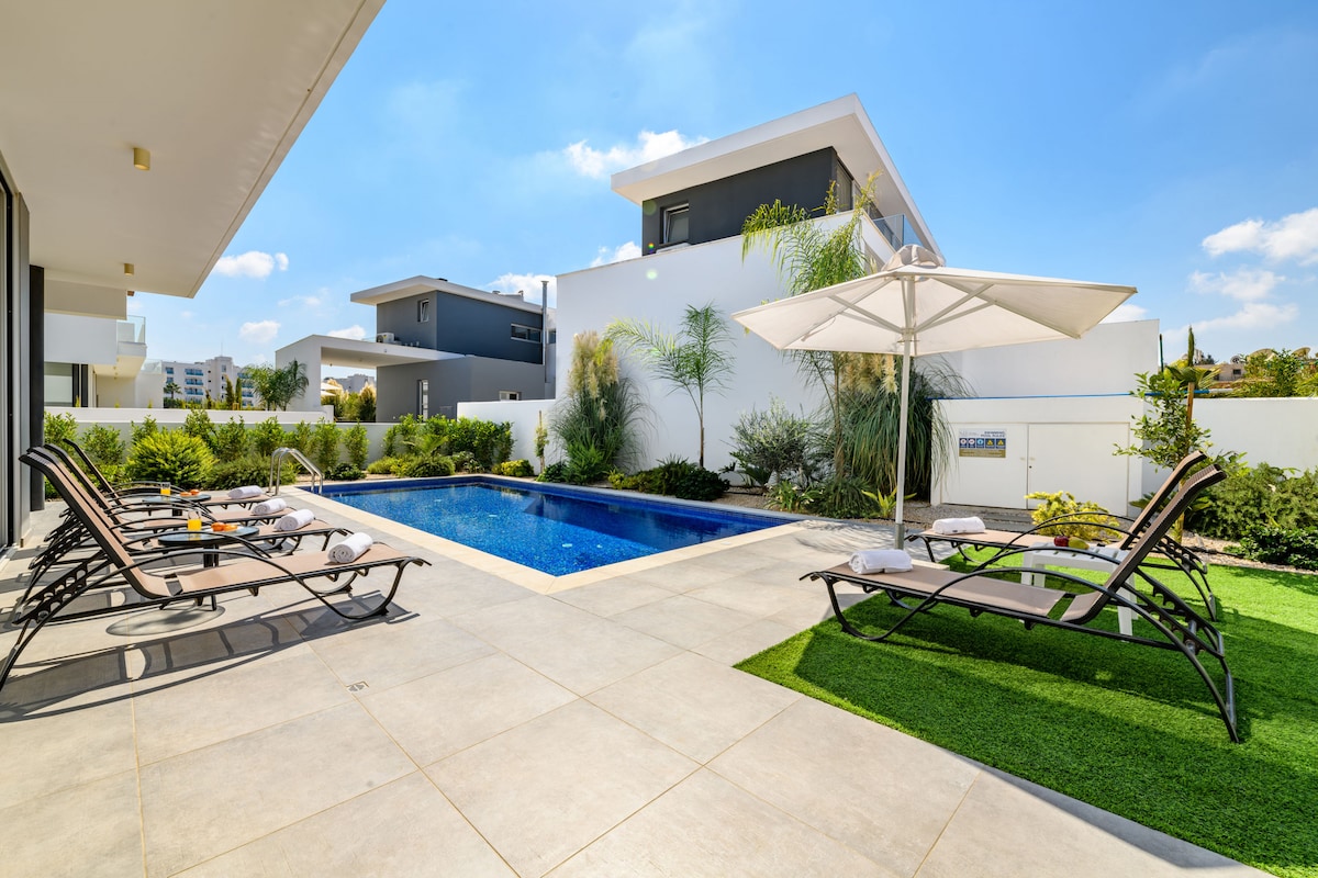 A serene outdoor area features a private swimming pool surrounded by loungers for sunbathing. An umbrella provides shade nearby, while landscaped greenery enhances the tranquil ambiance. The tiled patio offers ample space for relaxation and enjoying the outdoors.
