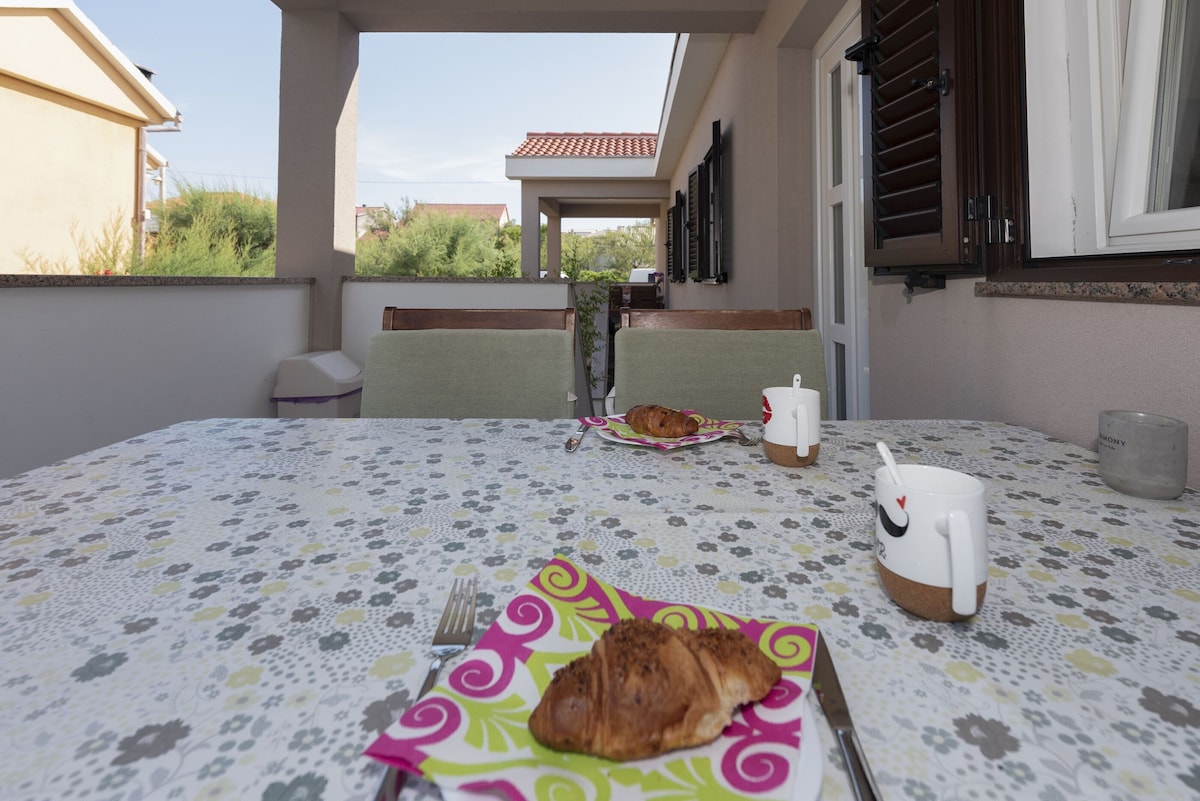 A dining area features a table set with colorful plates holding croissants and cups. The setting is outdoors, sheltered by a roof with light gray walls and shutters framing the space. Nearby greenery adds a natural touch to the relaxed atmosphere.