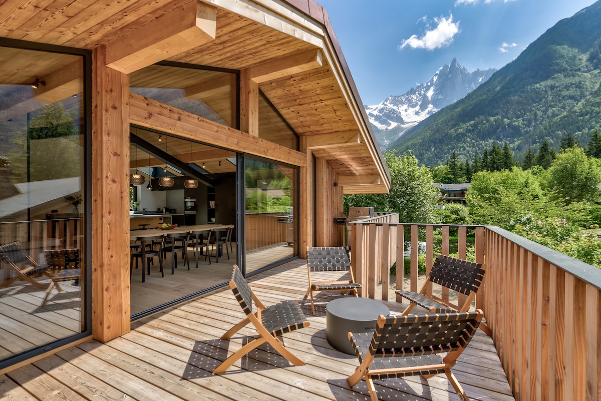 A spacious balcony features modern outdoor seating with wooden loungers arranged around a circular table. Floor-to-ceiling glass doors provide views into the open living area, while the backdrop showcases the mountainous landscape and clear blue sky.