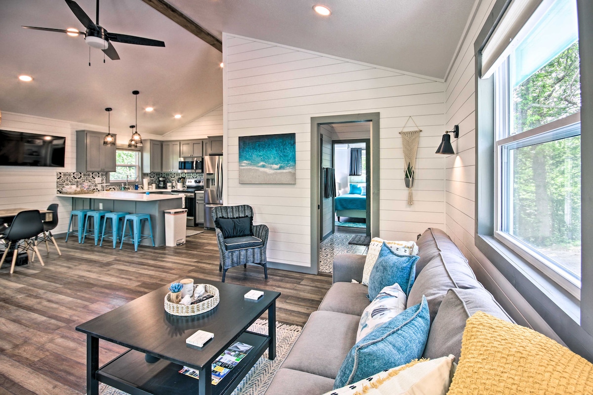 An open living area showcases a cozy seating arrangement with a coffee table, complemented by soft throw pillows. The modern kitchen, visible in the background, features a breakfast bar with blue stools. Bright windows invite natural light, enhancing the airy atmosphere.