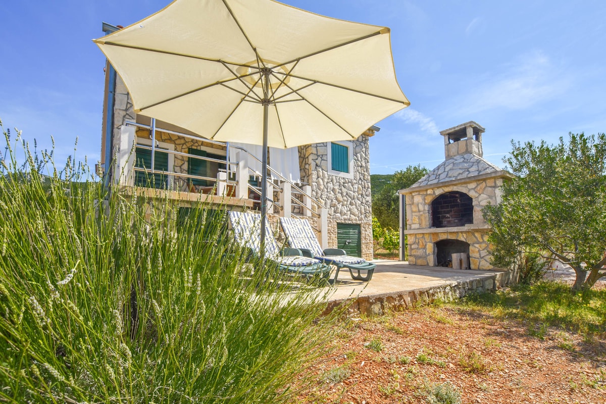 A patio area features a large umbrella providing shade over two lounge chairs. A stone barbecue is positioned nearby, while surrounding greenery and natural landscapes create a serene setting. The stone house with a green door is visible in the background.